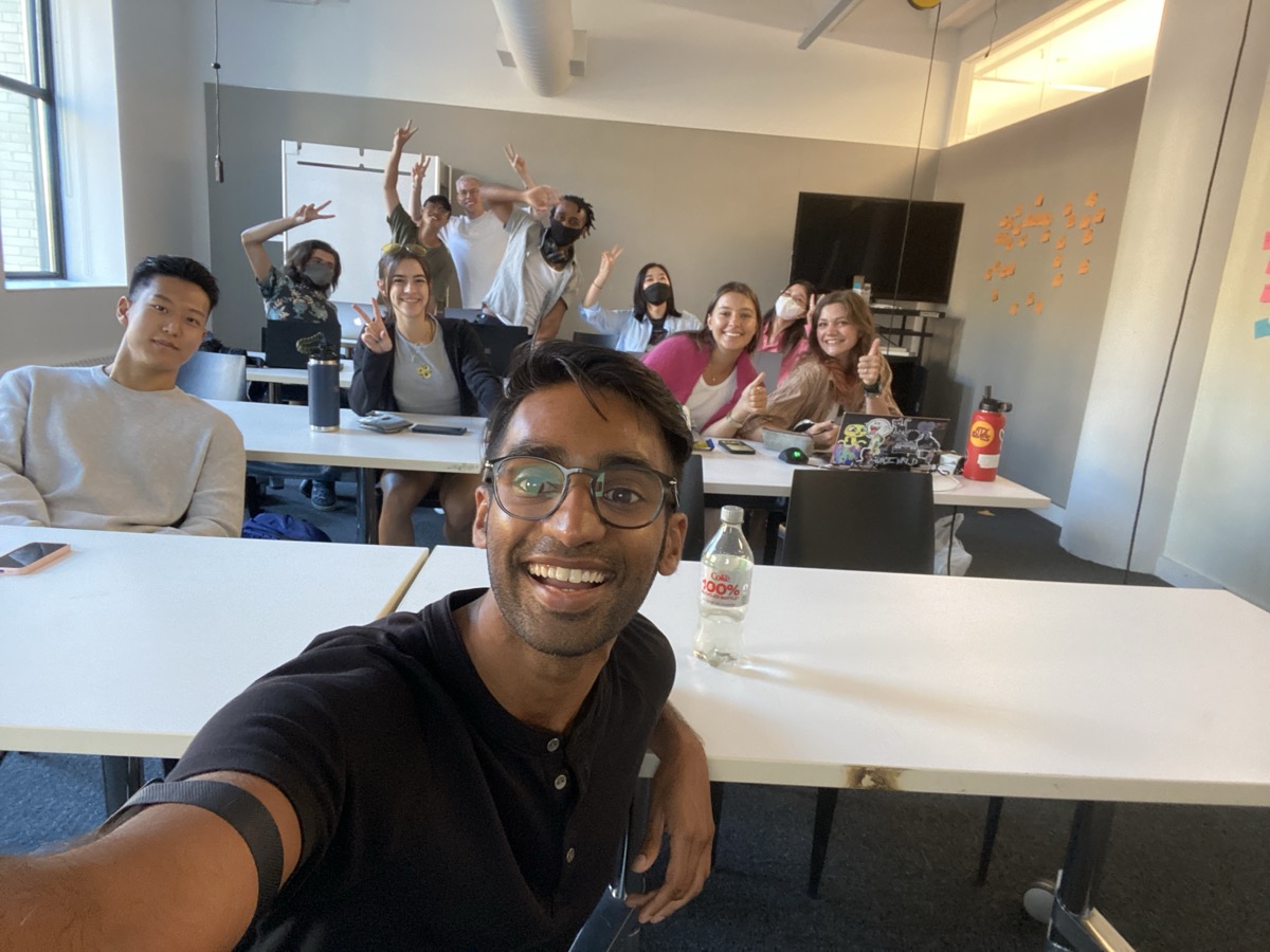 Jai holds a phone close for a classroom selfie while students smile, wave, and sit among laptops, water bottles, and sticky notes.